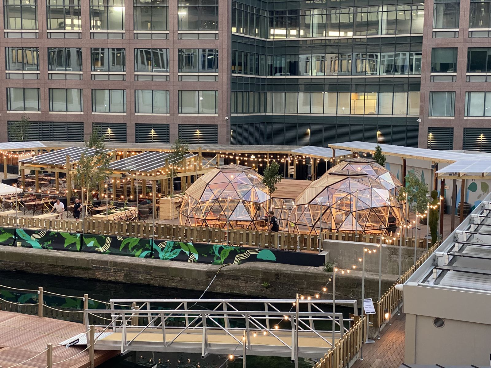 Three cedar and glazed Wilder domes glowing at dusk on the Eden Dock terrace, Canary Wharf towers behind